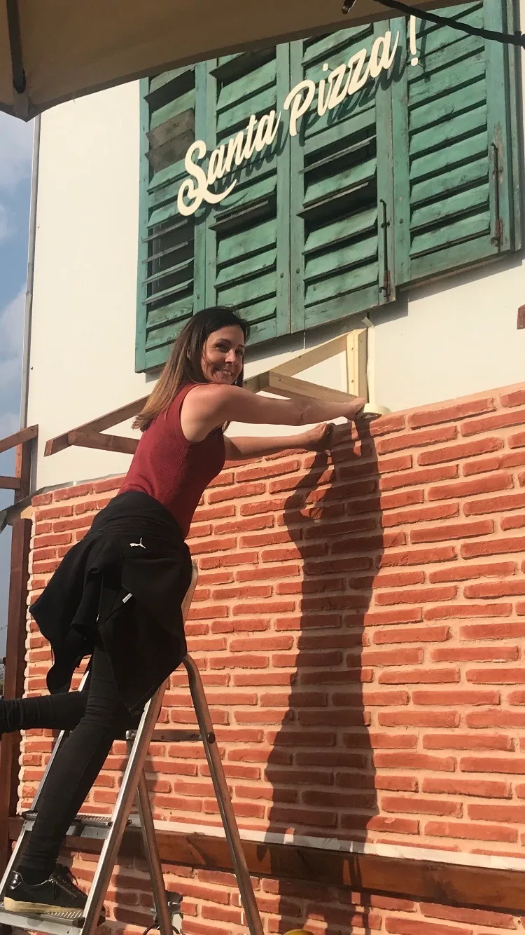 Eine Frau steht auf einer Leiter und bringt ein Klebeband an der Wand an. Im Hintergrund steht Santa Pizza auf grünen Fensterläden.